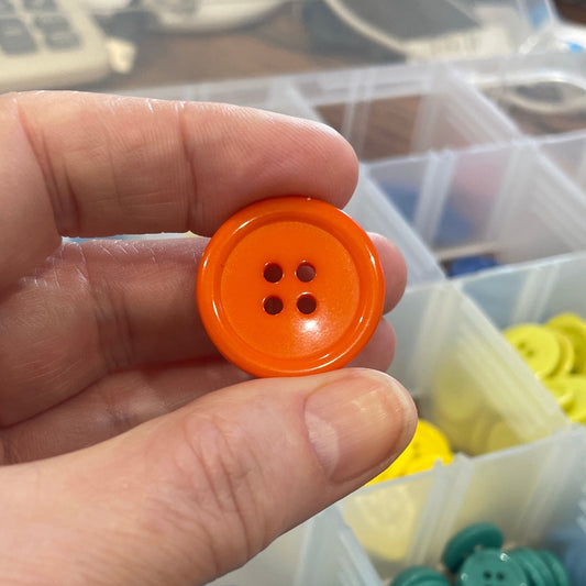 Hand holding an orange button with a container of colorful buttons in the background.