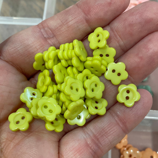 Hand holding small yellow flower-shaped beads with a container of more beads in the background.