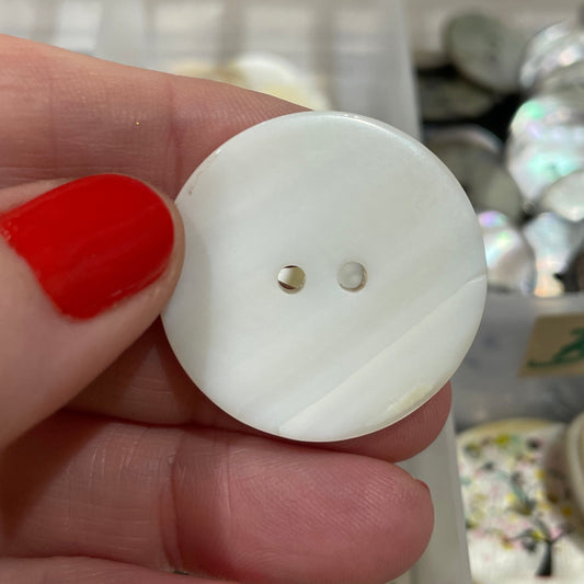 Hand holding a white button with a small hole, with a blurred background of buttons and containers.