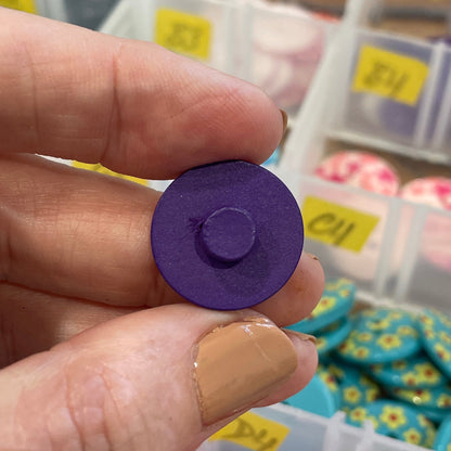 Hand holding a purple button with a container of buttons in the background