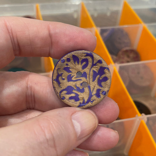Hand holding a decorative button with intricate designs, against a background of a button organizer.