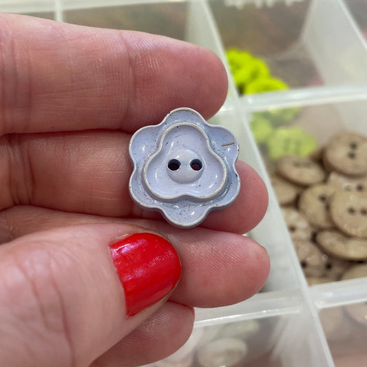 Hand holding a white button with a flower design over a container of various buttons.