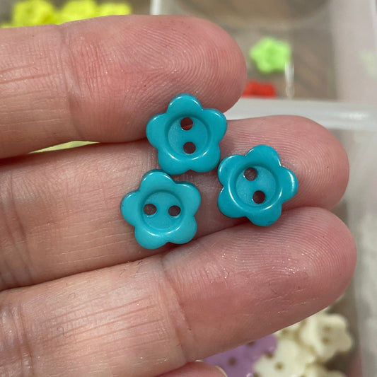 Turquoise flower-shaped buttons held between fingers with a blurred background of colorful buttons.