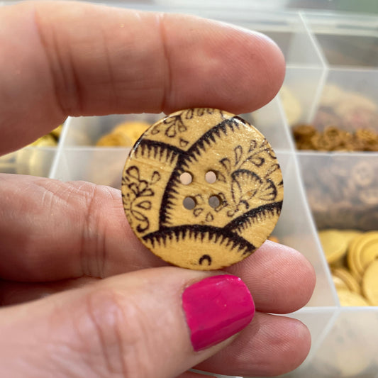 Hand holding a decorative wooden button with a detailed design, against a background of various buttons.