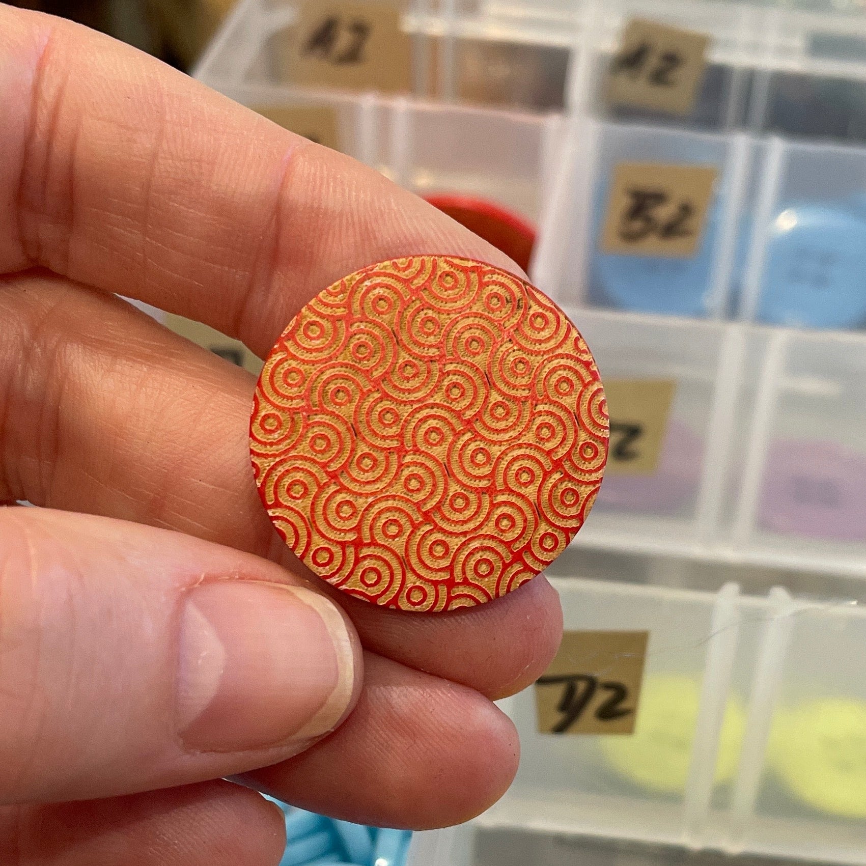 Hand holding a button, against a background of a clear plastic container with compartments.