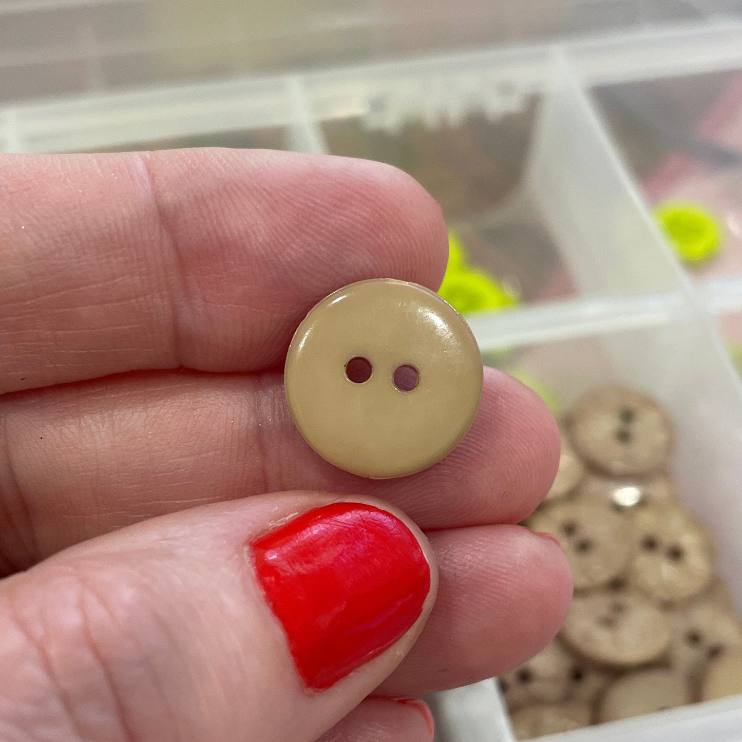 Hand holding a beige button with a container of various buttons in the background