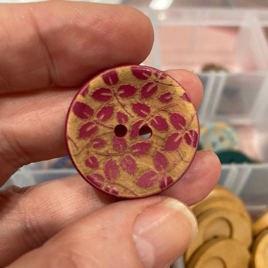 Hand holding a wooden button with floral pattern in front of a container of buttons.