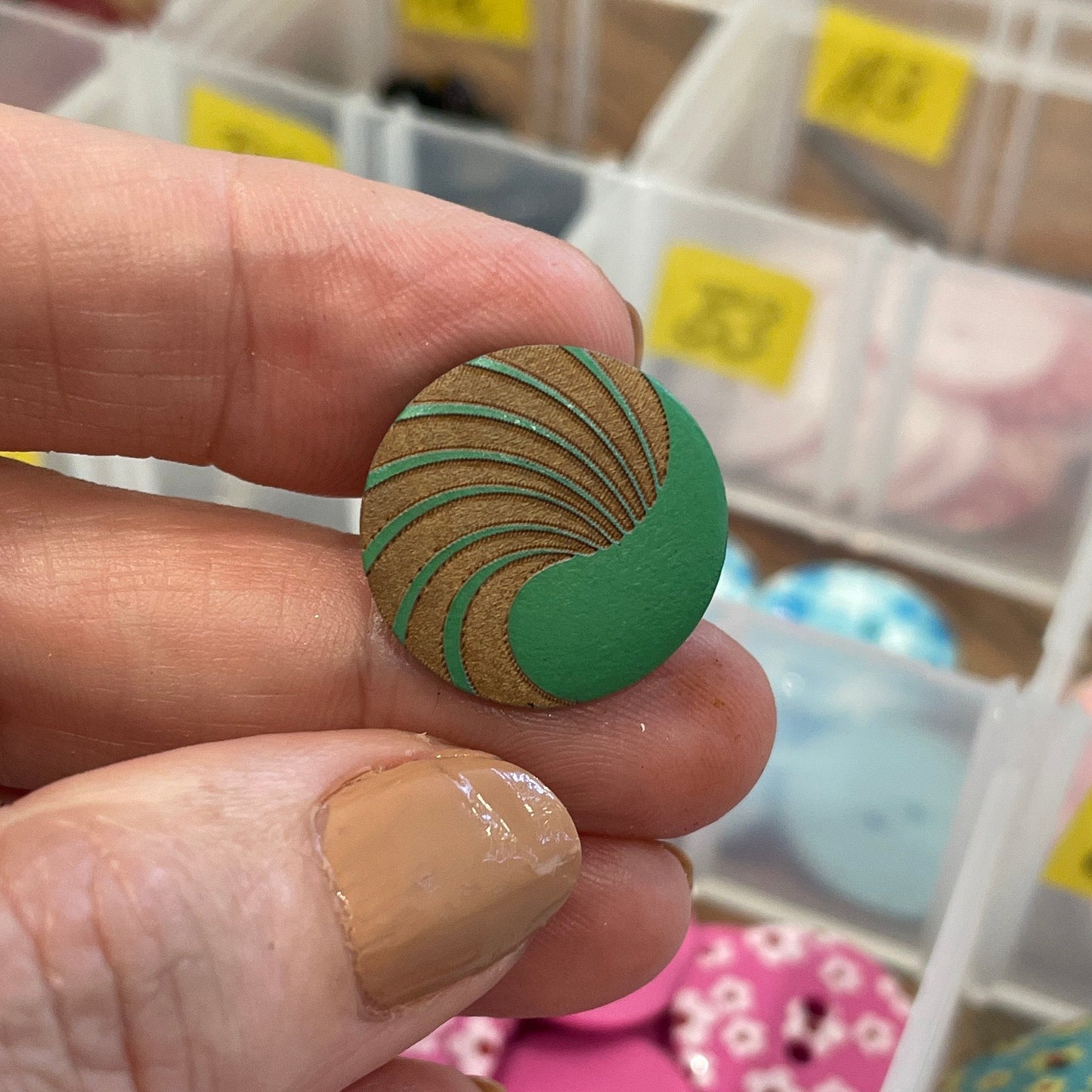 Hand holding a green button with a pattern, surrounded by various buttons in a container.