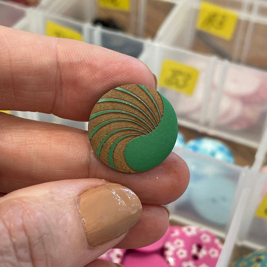 Hand holding a green button with a pattern, surrounded by various buttons in a container.