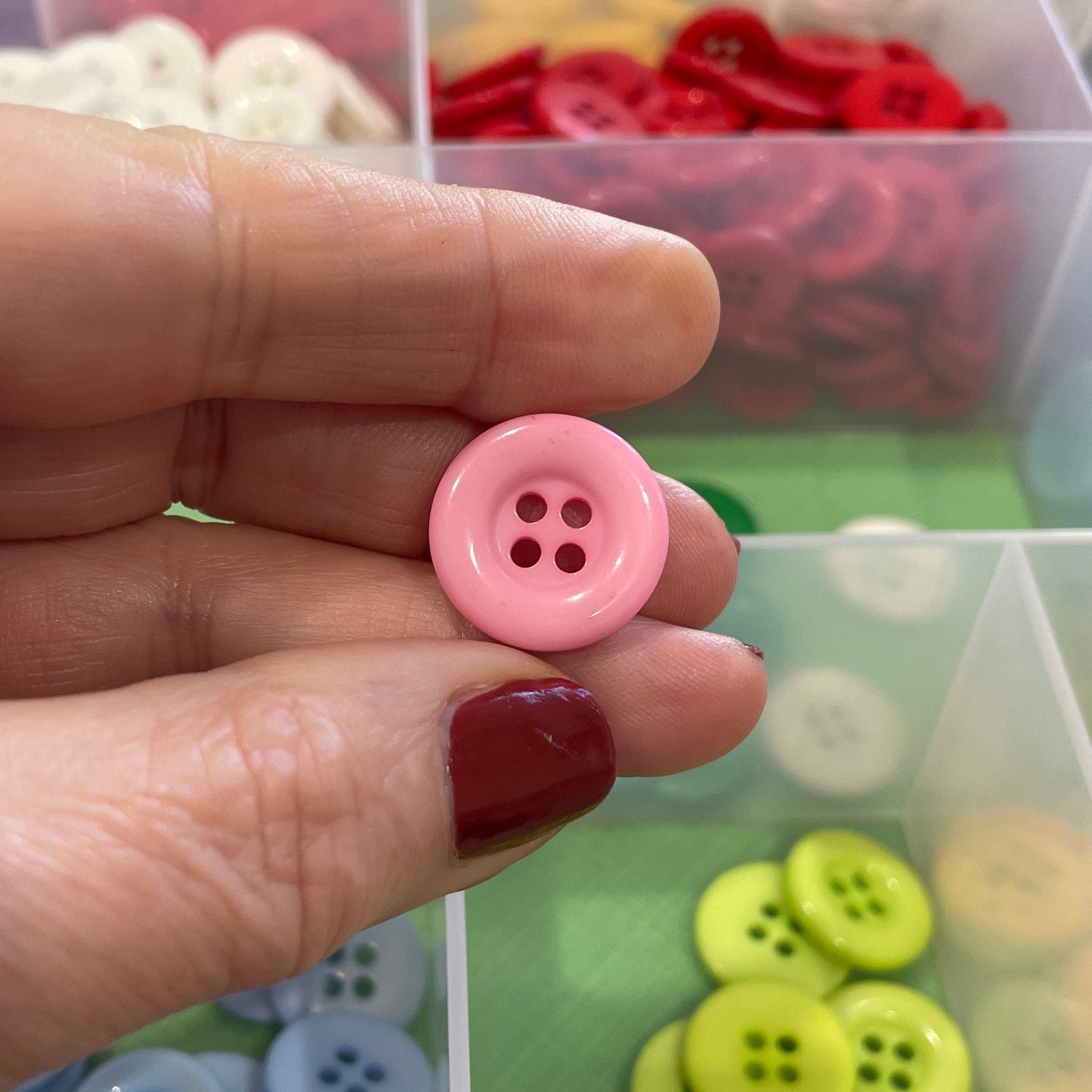 Hand holding a pink button with a container of assorted buttons in the background