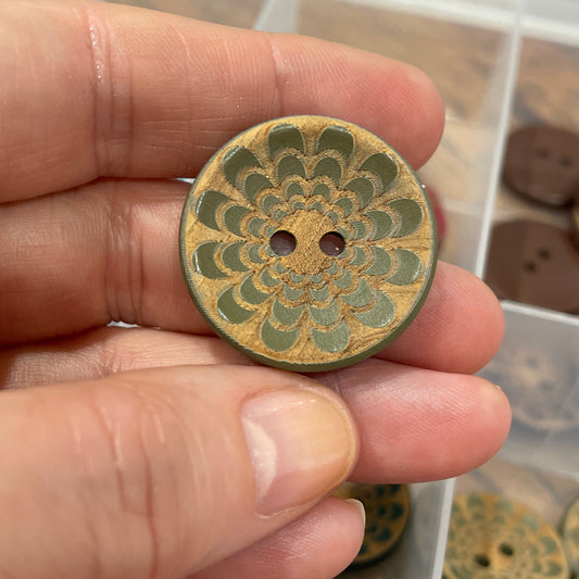 Hand holding a decorative button with intricate design, set against a background of similar buttons in a box.