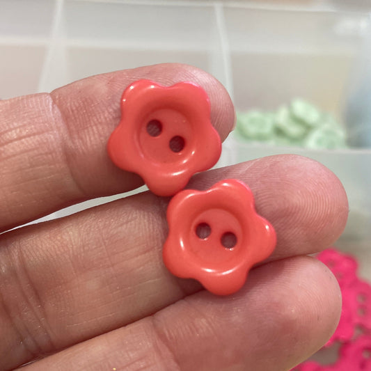 Hand holding two pink flower-shaped buttons with a blurred background of colorful buttons.