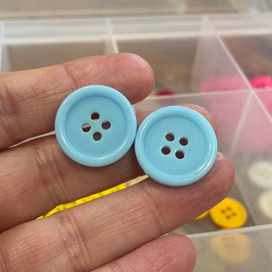 Hand holding two blue buttons with a container of colorful buttons in the background