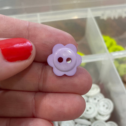 Hand holding a small purple flower-shaped button with a clear container of buttons in the background.