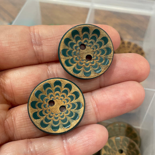 Hand holding two decorative wooden buttons with intricate patterns, with a display box of more buttons in the background.
