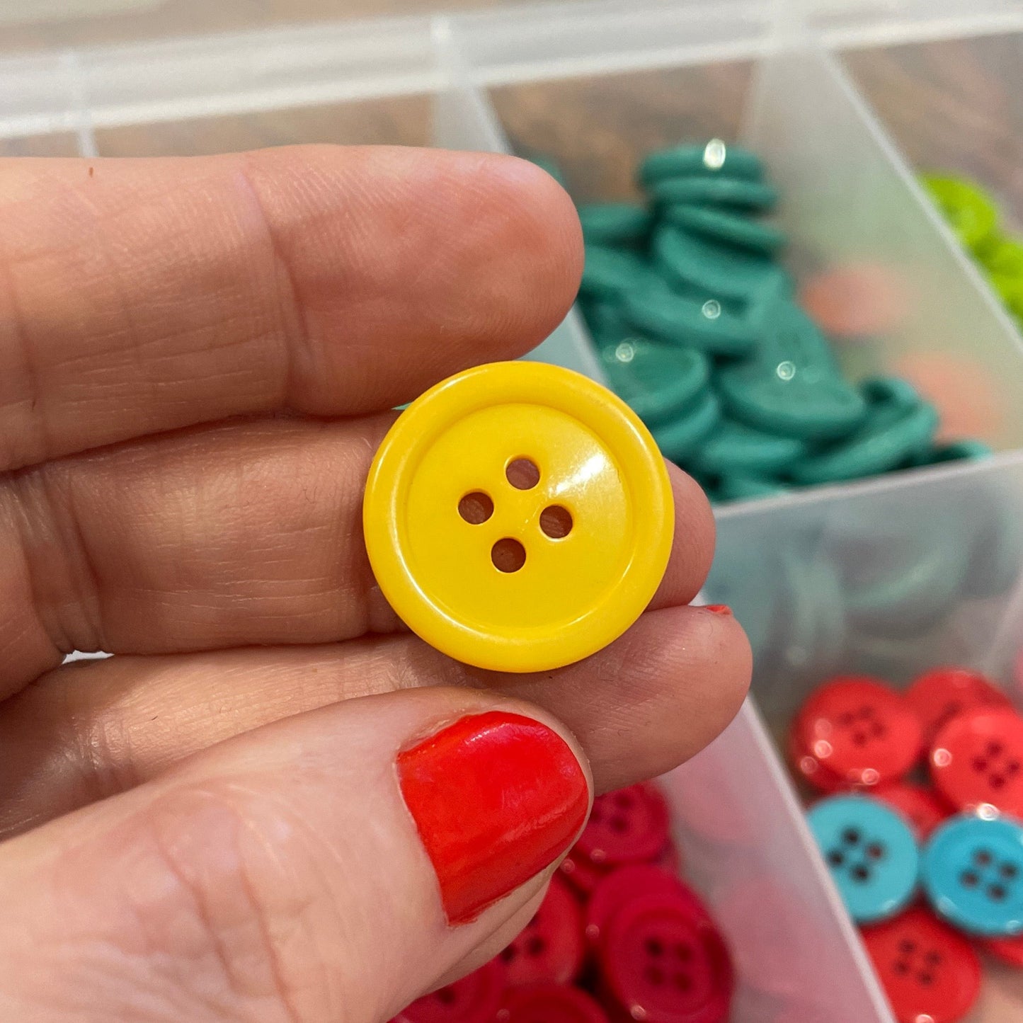 Hand holding a yellow button with red nail polish, surrounded by colorful buttons in a clear container.