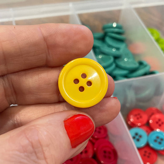 Hand holding a yellow button with red nail polish, surrounded by colorful buttons in a clear container.
