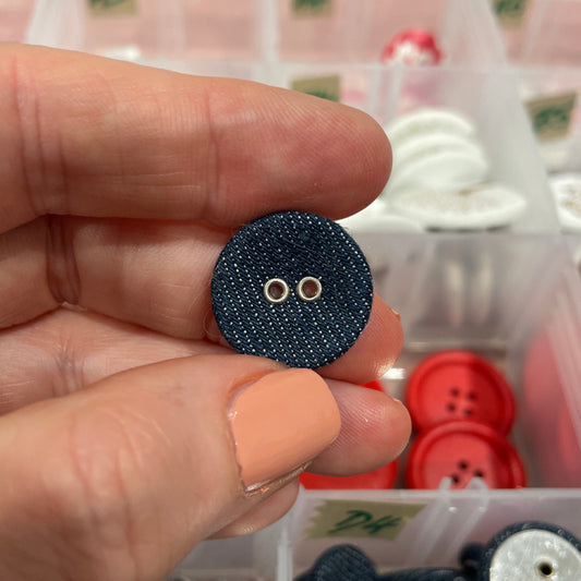 Hand holding a blue button with a background of various buttons in containers.