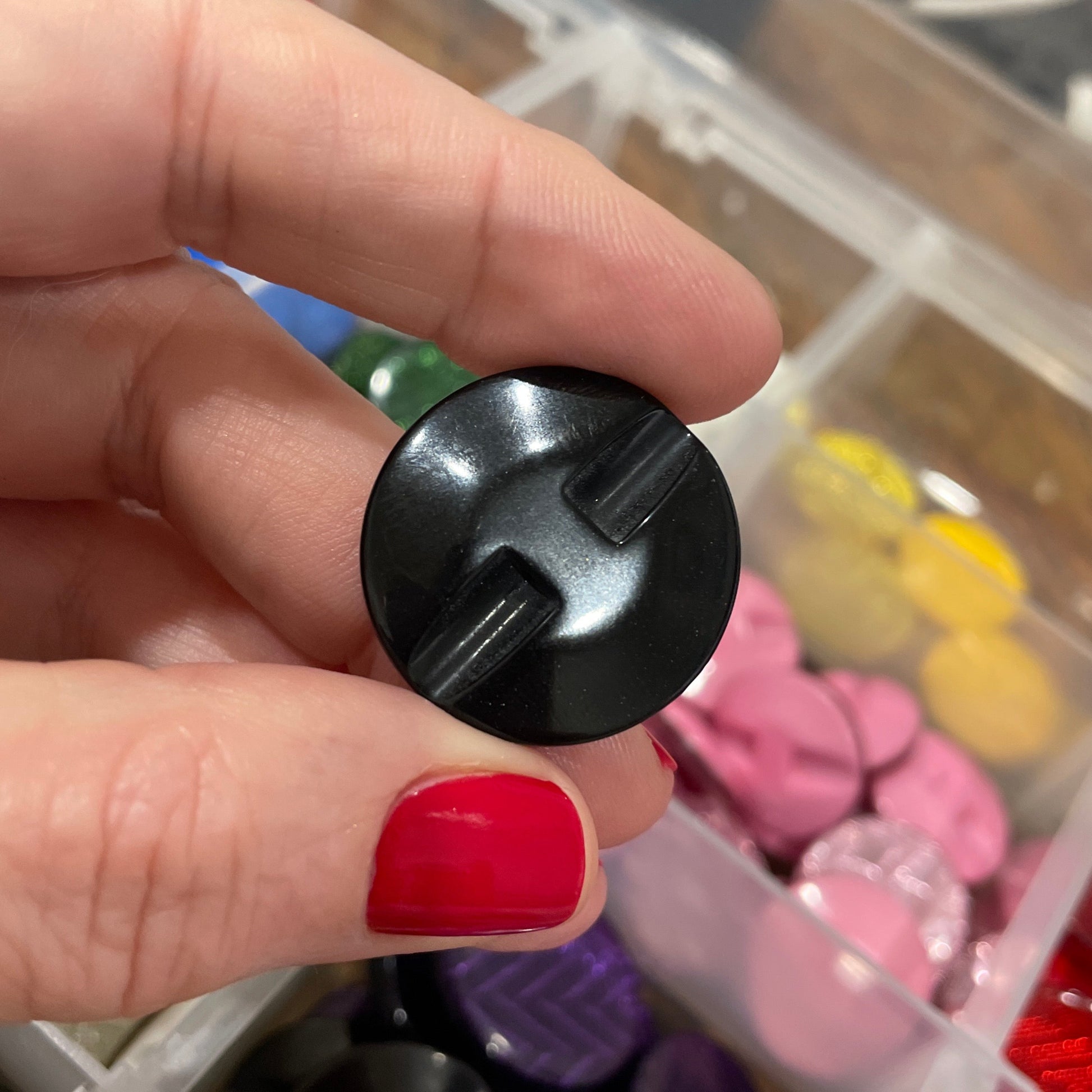 Black plastic component held by a hand with red nail polish, against a background of colorful plastic items.