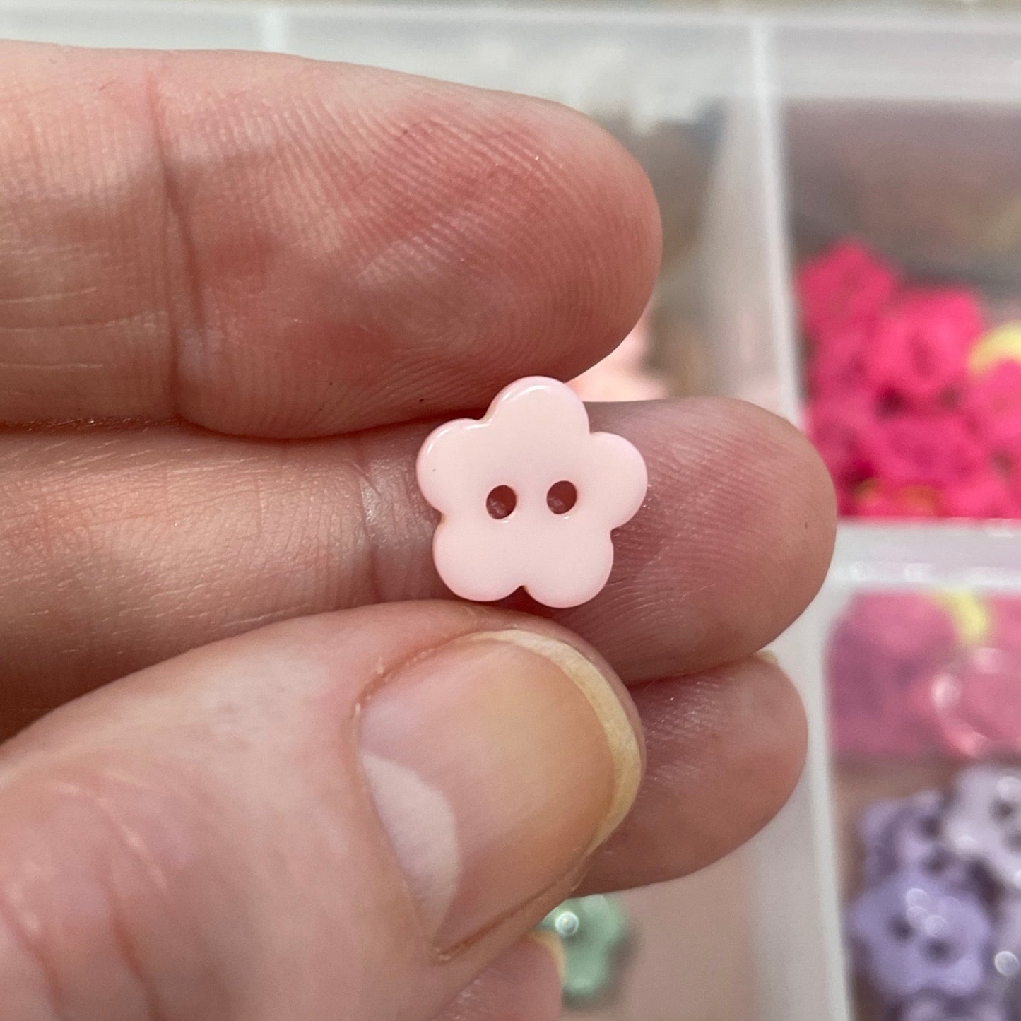 Hand holding a small pink flower-shaped object with a container of colorful buttons in the background.