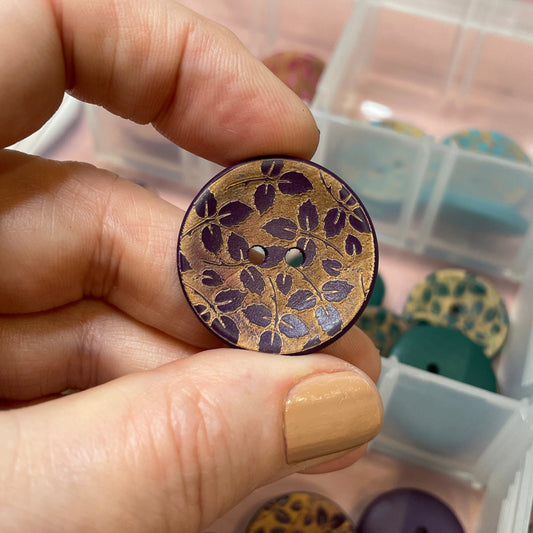Hand holding a decorative button with a leopard print pattern, surrounded by more buttons in a container.