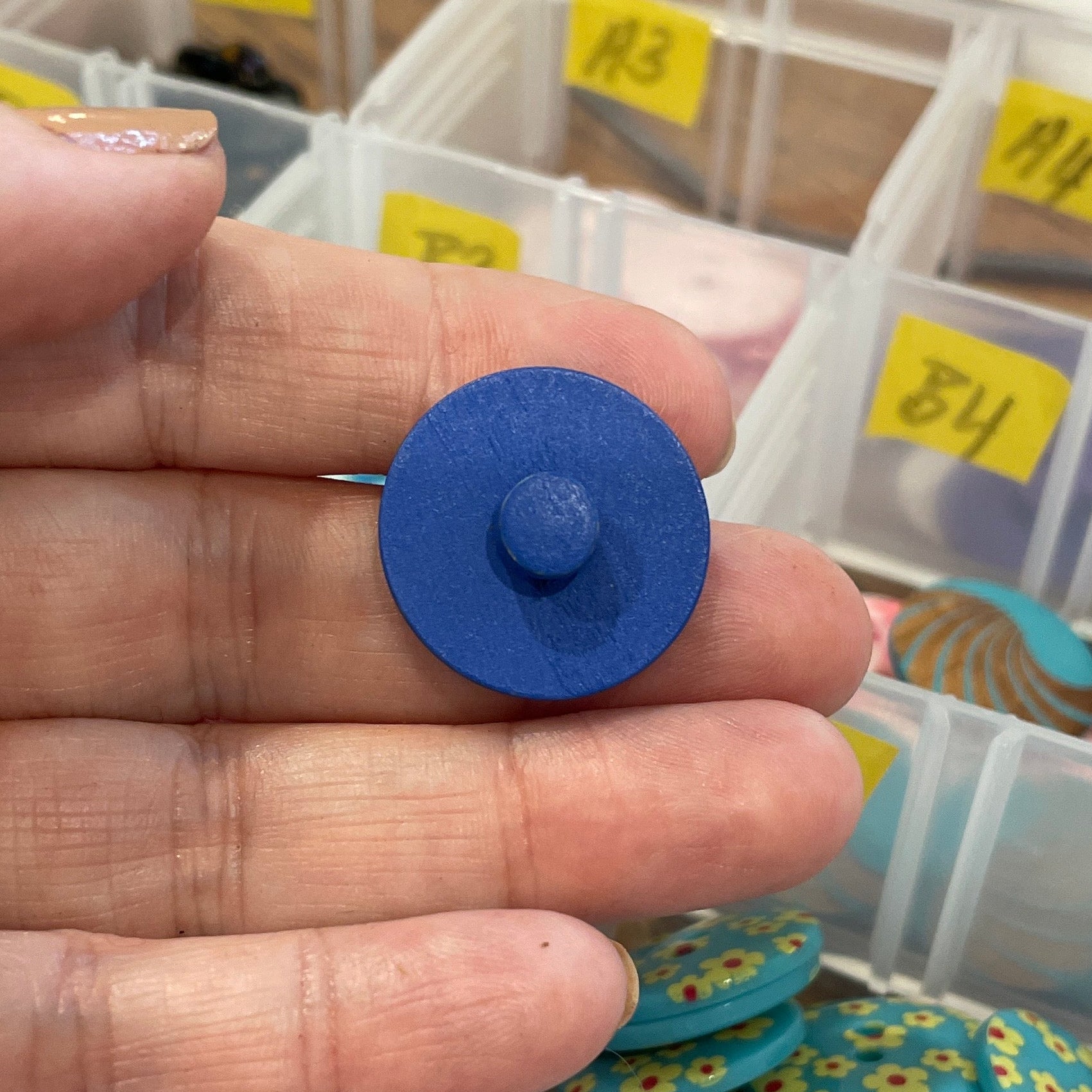 Hand holding a blue button with a container of various buttons in the background