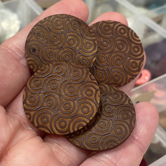 Hand holding decorative wooden buttons with intricate patterns against a blurred background of a storage container.