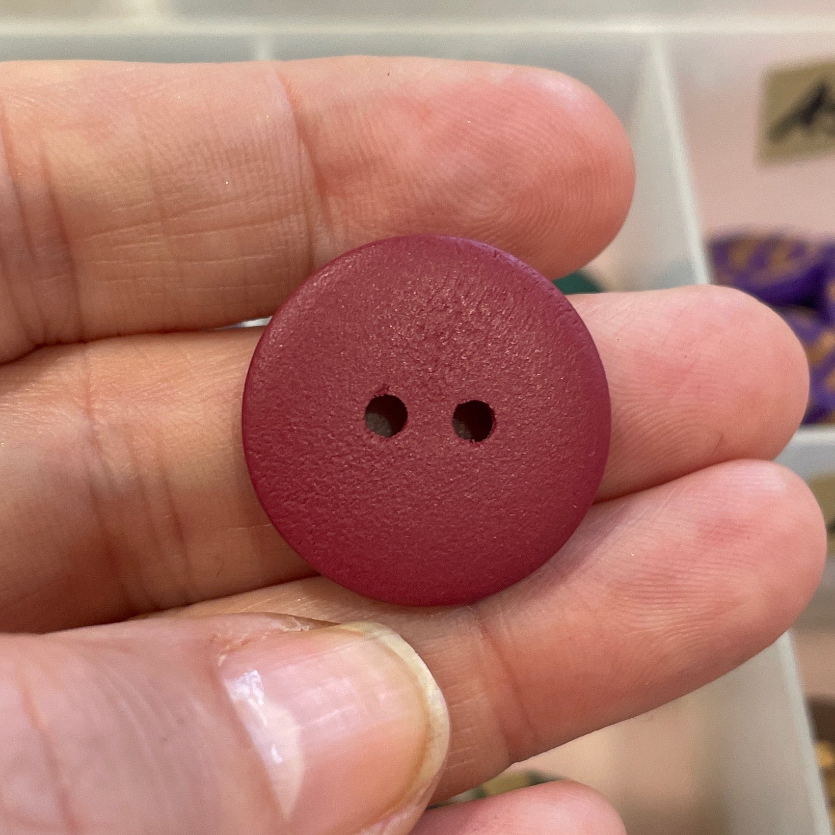 Maroon button held in a hand with a background of various buttons in a container.