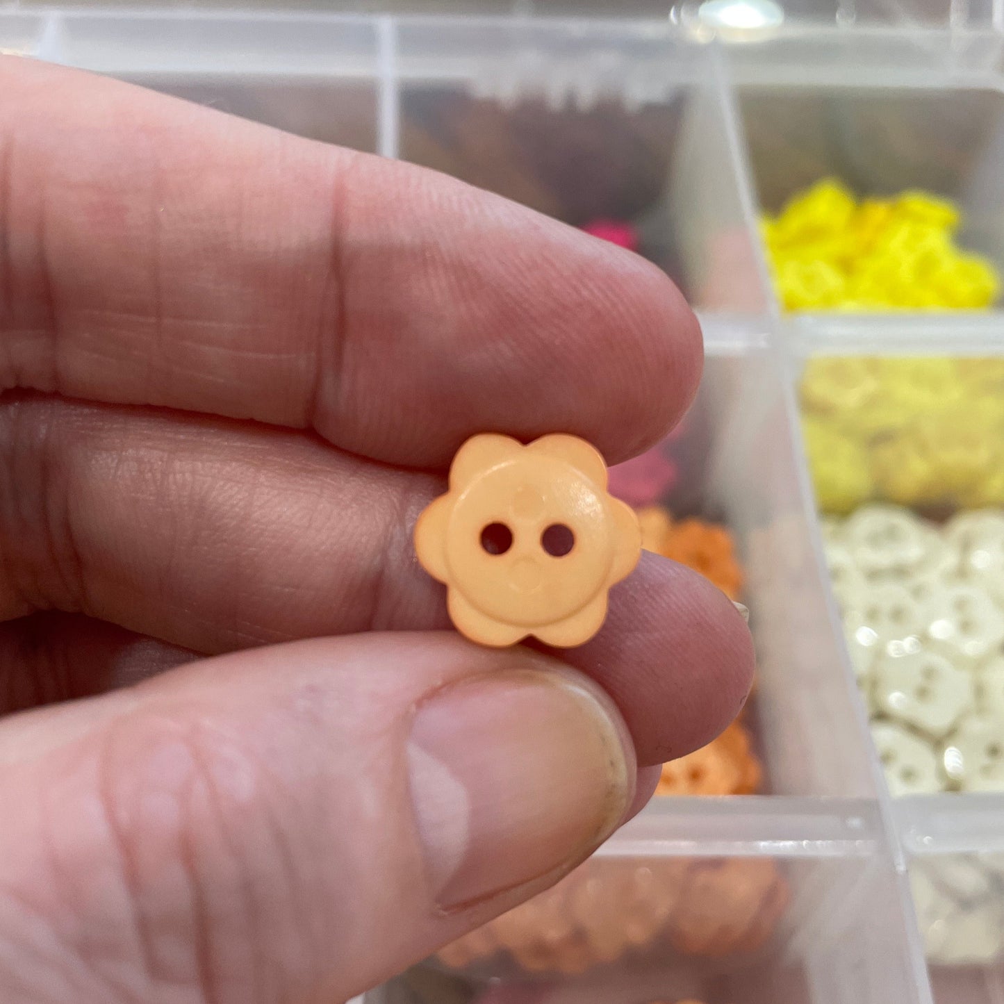 Hand holding a small yellow flower-shaped button with a blurred container of buttons in the background.