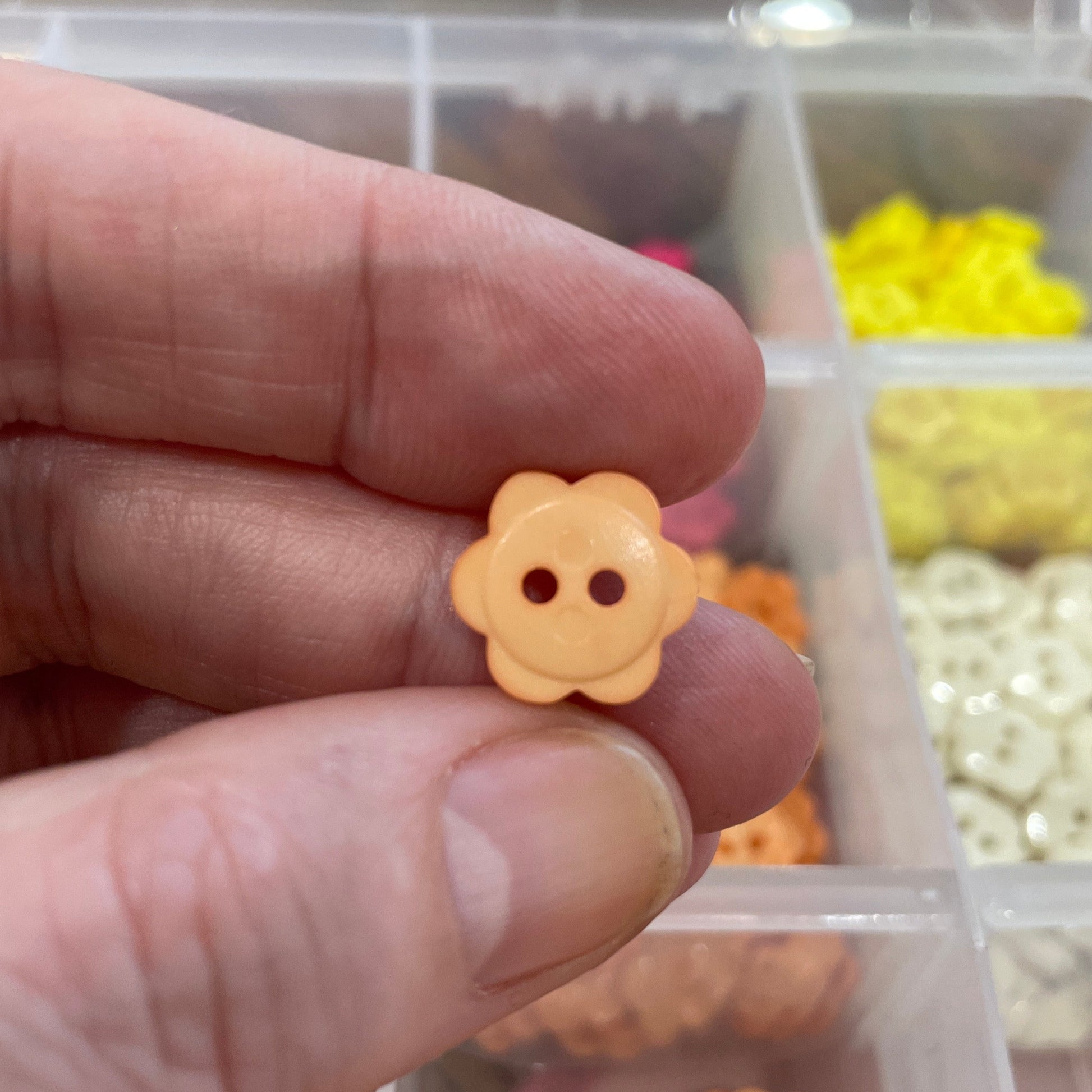 Hand holding a small yellow flower-shaped button with a blurred container of buttons in the background.