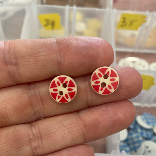 Hand holding two red and white buttons with a pattern, with a container of buttons in the background.