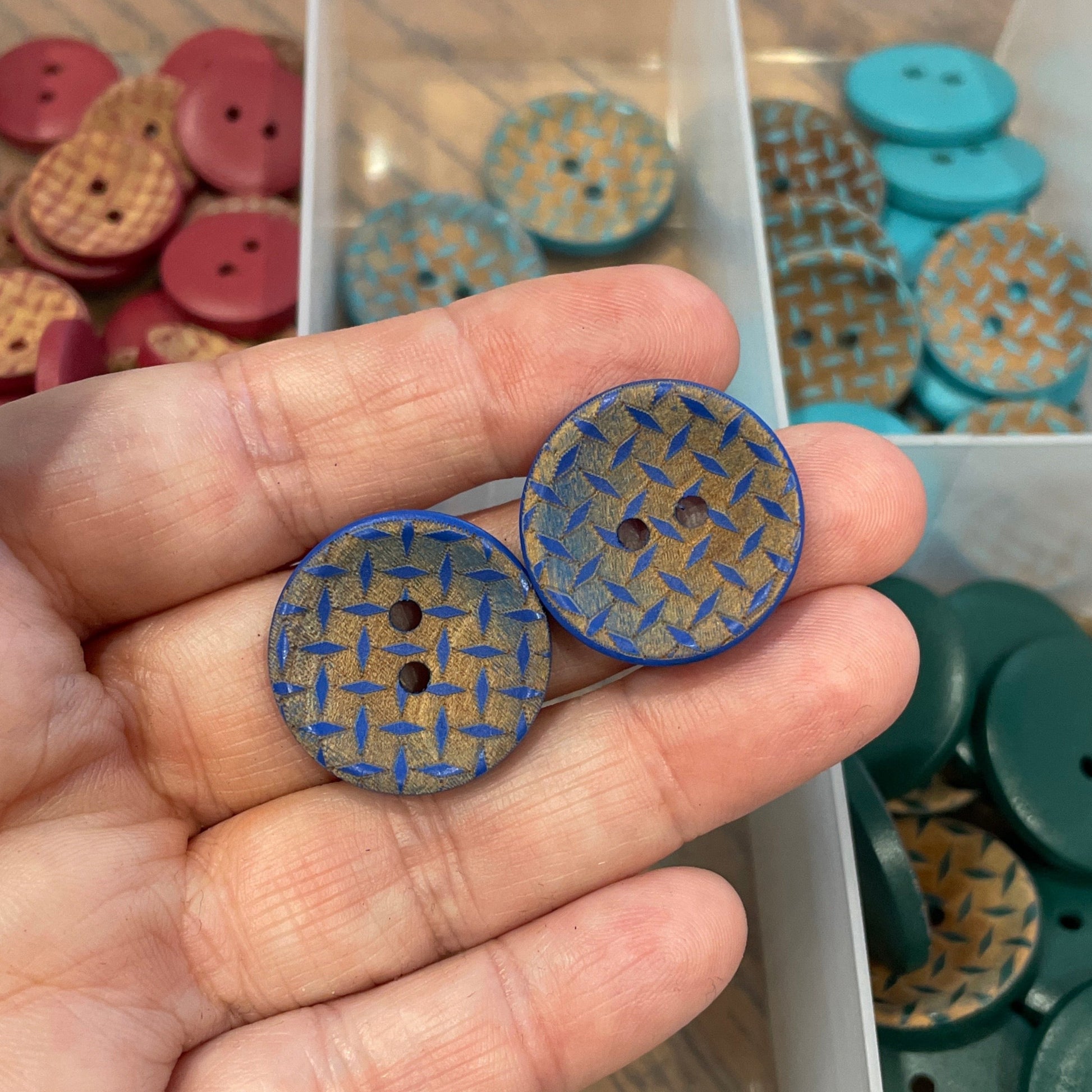 Hand holding two blue buttons with a container of assorted buttons in the background
