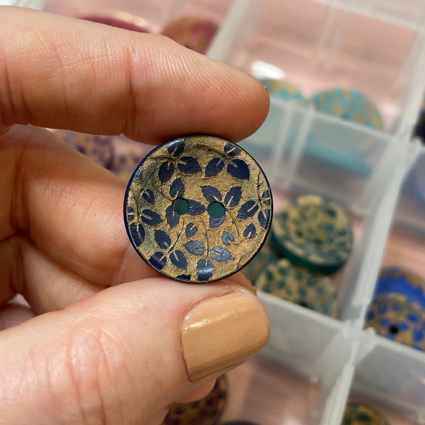 Hand holding a decorative button with a floral pattern over a container of buttons.