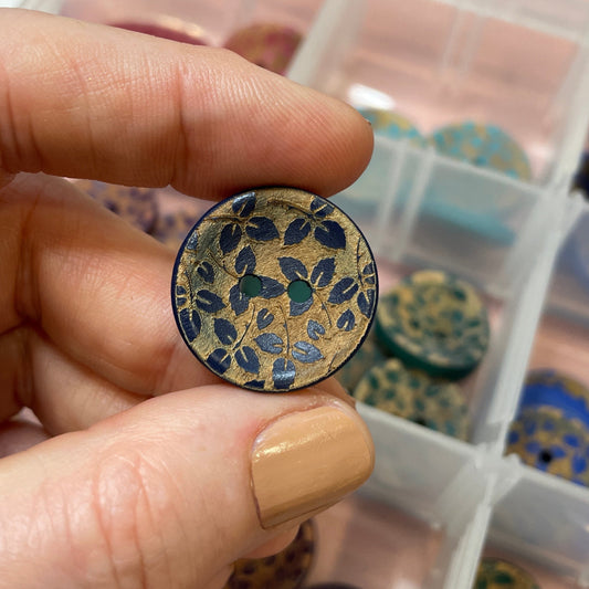 Hand holding a decorative button with a floral pattern over a container of buttons.