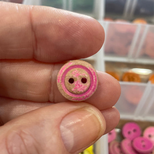 Hand holding a pink button with a blurred container of buttons in the background