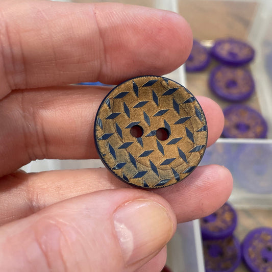 Hand holding a decorative button with a geometric pattern, against a background of various buttons in a container.