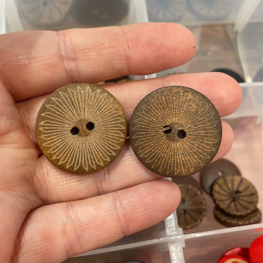 Hand holding two brown buttons with a clear container of assorted buttons in the background