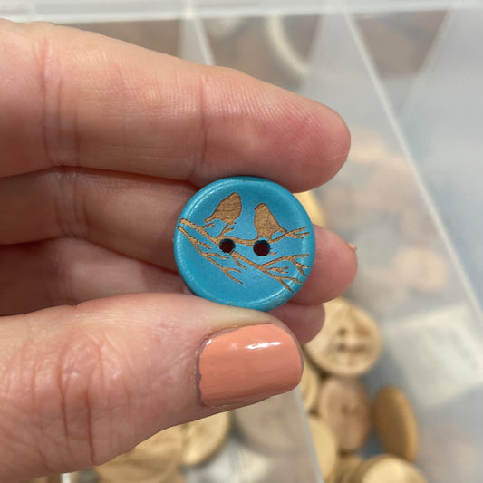 Hand holding a blue button with gold design in front of a container of wooden buttons.