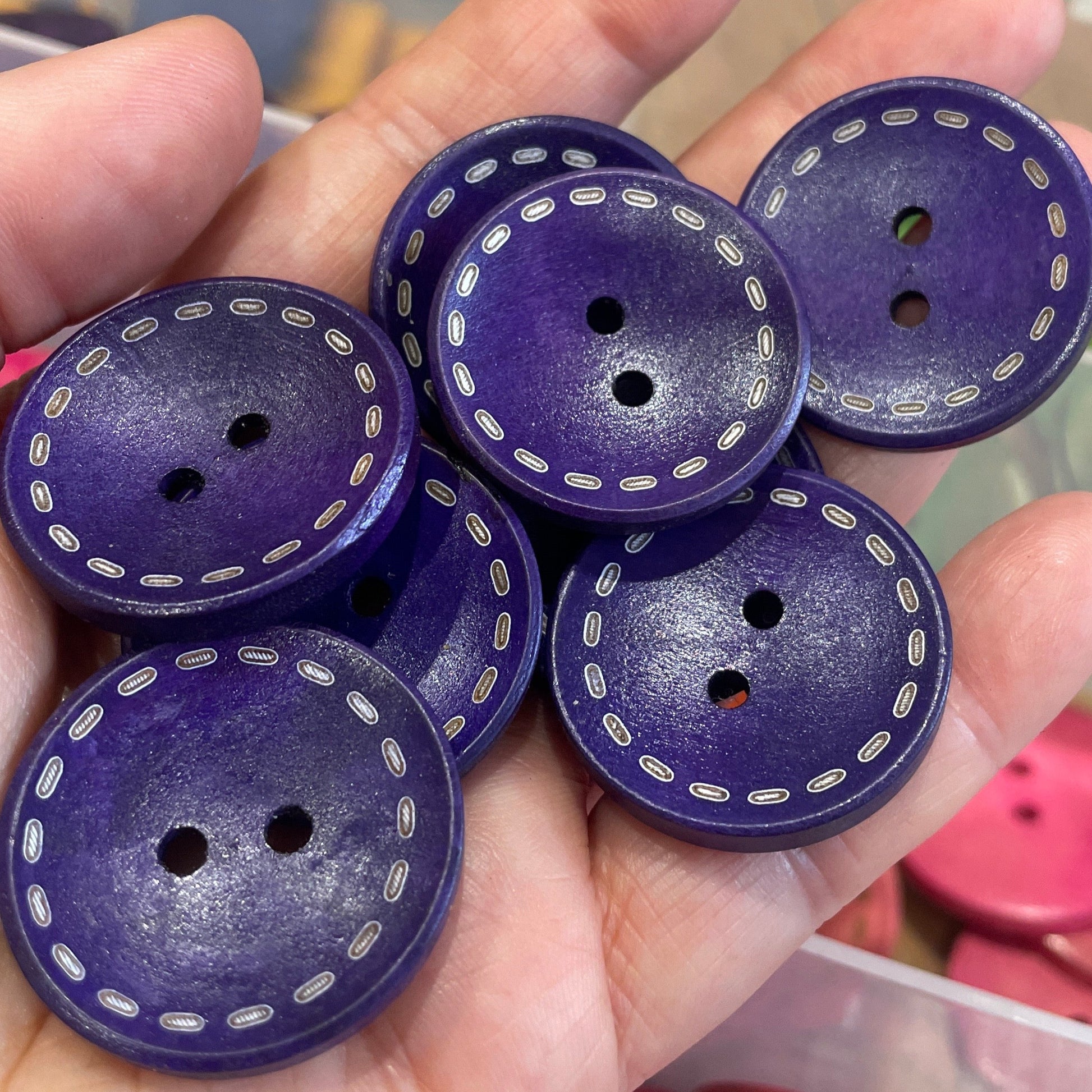 Purple buttons with white stitching held in a hand, with a blurred background of pink buttons and a container.