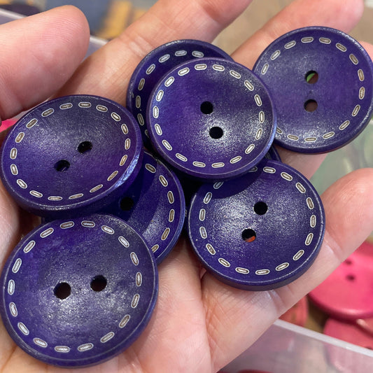 Purple buttons with white stitching held in a hand, with a blurred background of pink buttons and a container.