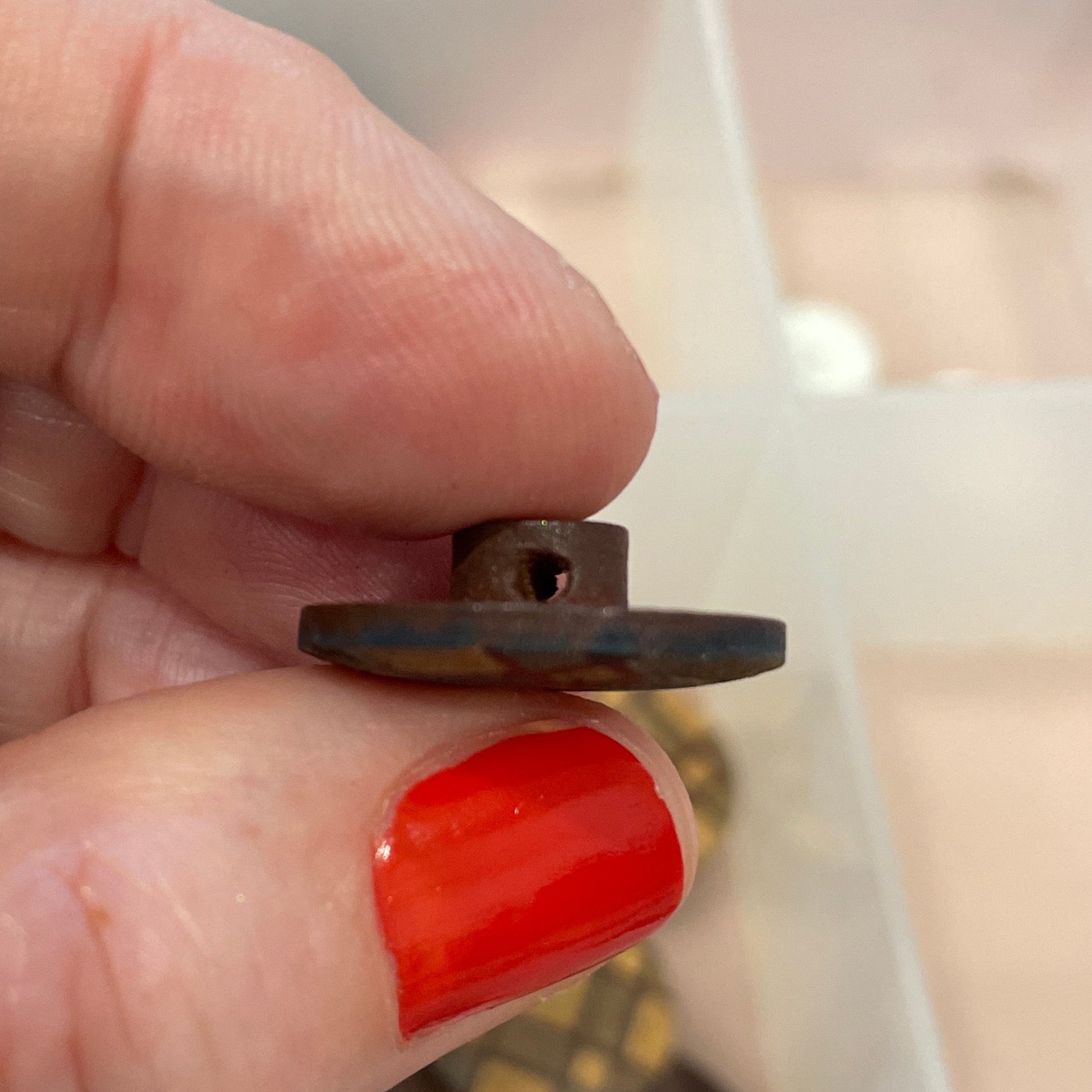 Hand holding a small metal object with a clear container of buttons in the background