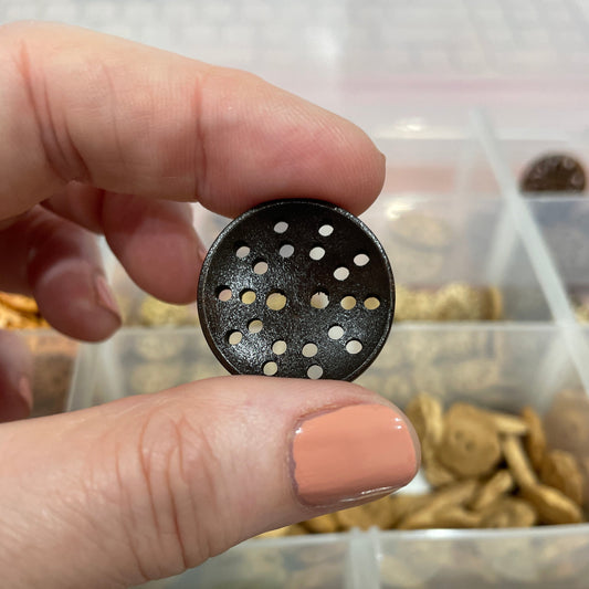 Hand holding a button with a unique design, with a container of buttons in the background.