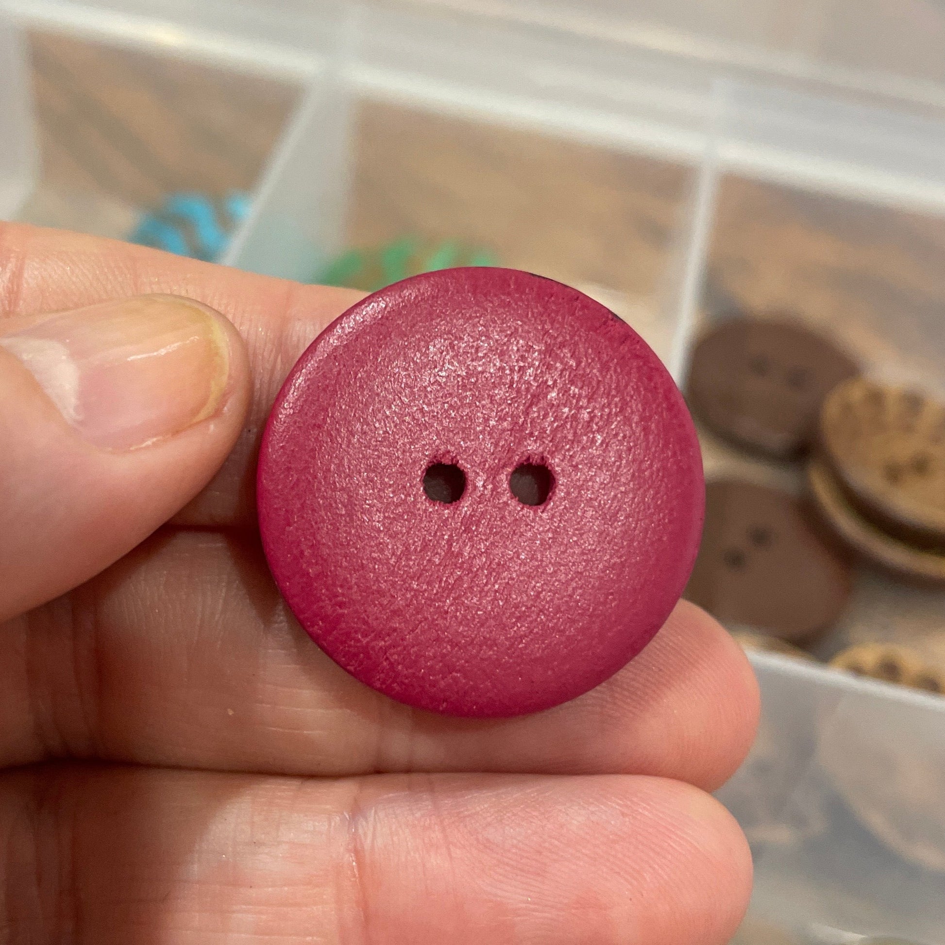 Hand holding a pink button with a blurred container of buttons in the background