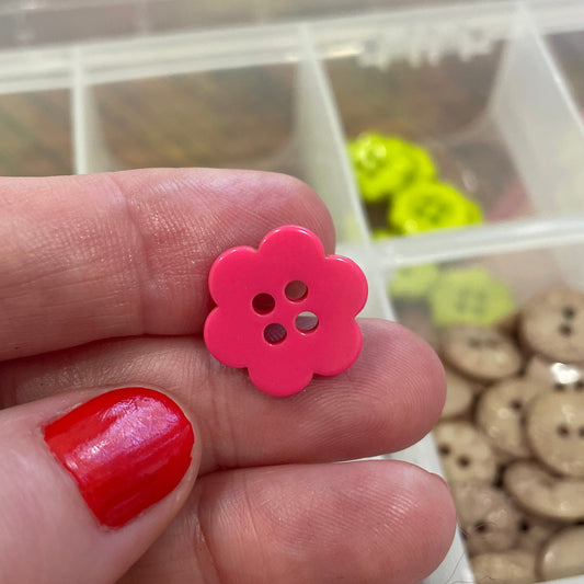 Hand holding a pink flower-shaped button with a container of various buttons in the background.