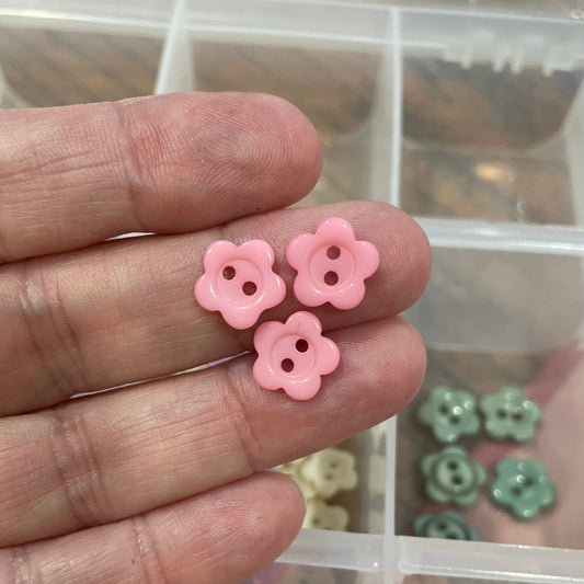Hand holding pink flower-shaped buttons with a container of similar buttons in the background.