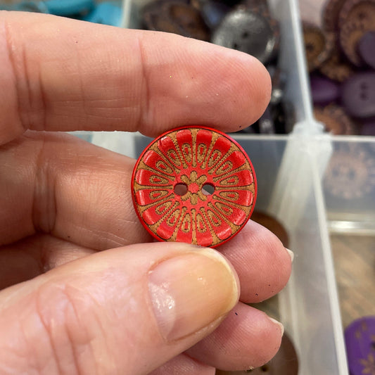 Hand holding a red button with intricate design in front of a container of various buttons.