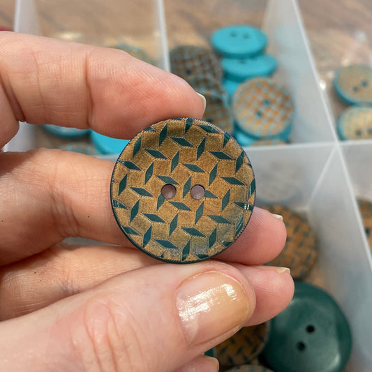 Hand holding a patterned button with a container of buttons in the background