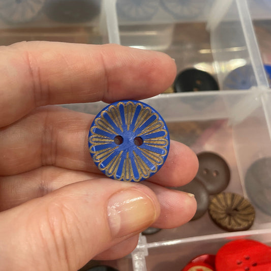 Hand holding a blue decorative button with a clear container of assorted buttons in the background.