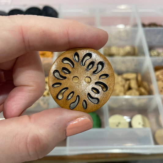 Hand holding a decorative wooden button with a cut-out design against a blurred background of a button collection.