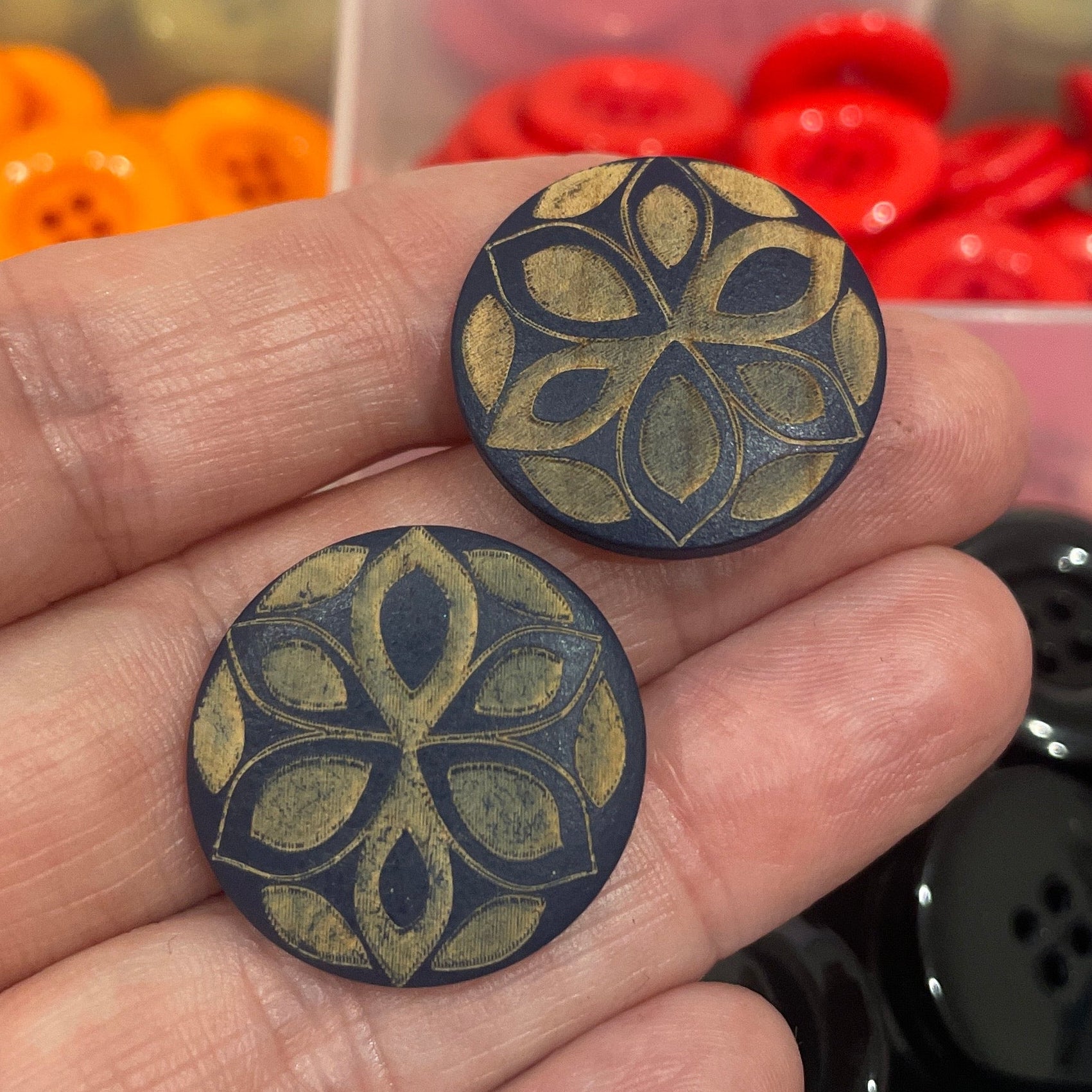 Hand holding two decorative buttons with a background of assorted buttons in a box.
